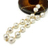 A strand necklace made of irregular, hammered white pearls connected by delicate gold chains, displayed on a clean white background with a dark wooden stand partially visible in the upper right.