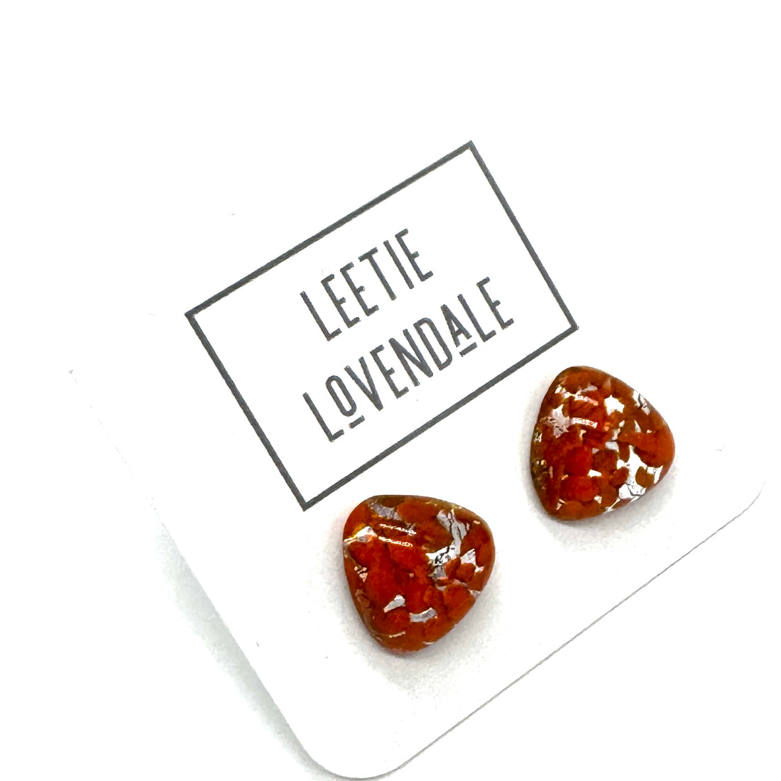 Two small triangular-shaped clear glass stud earrings with orange inclusions, displayed on a white card printed with the name &