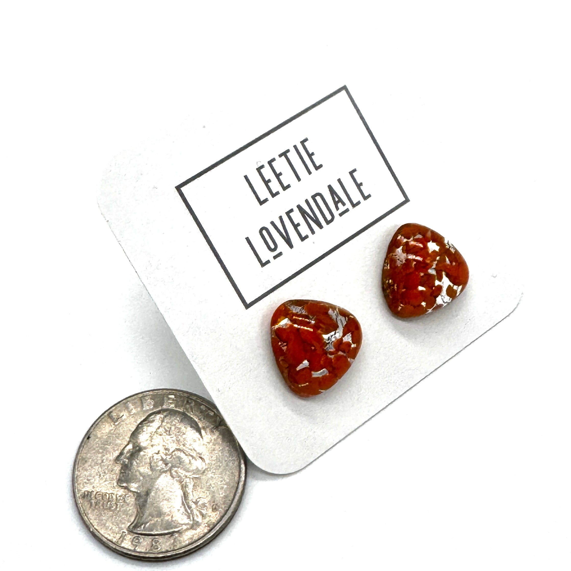 Two small triangular-shaped clear glass stud earrings with orange inclusions, displayed on a white card printed with the name &