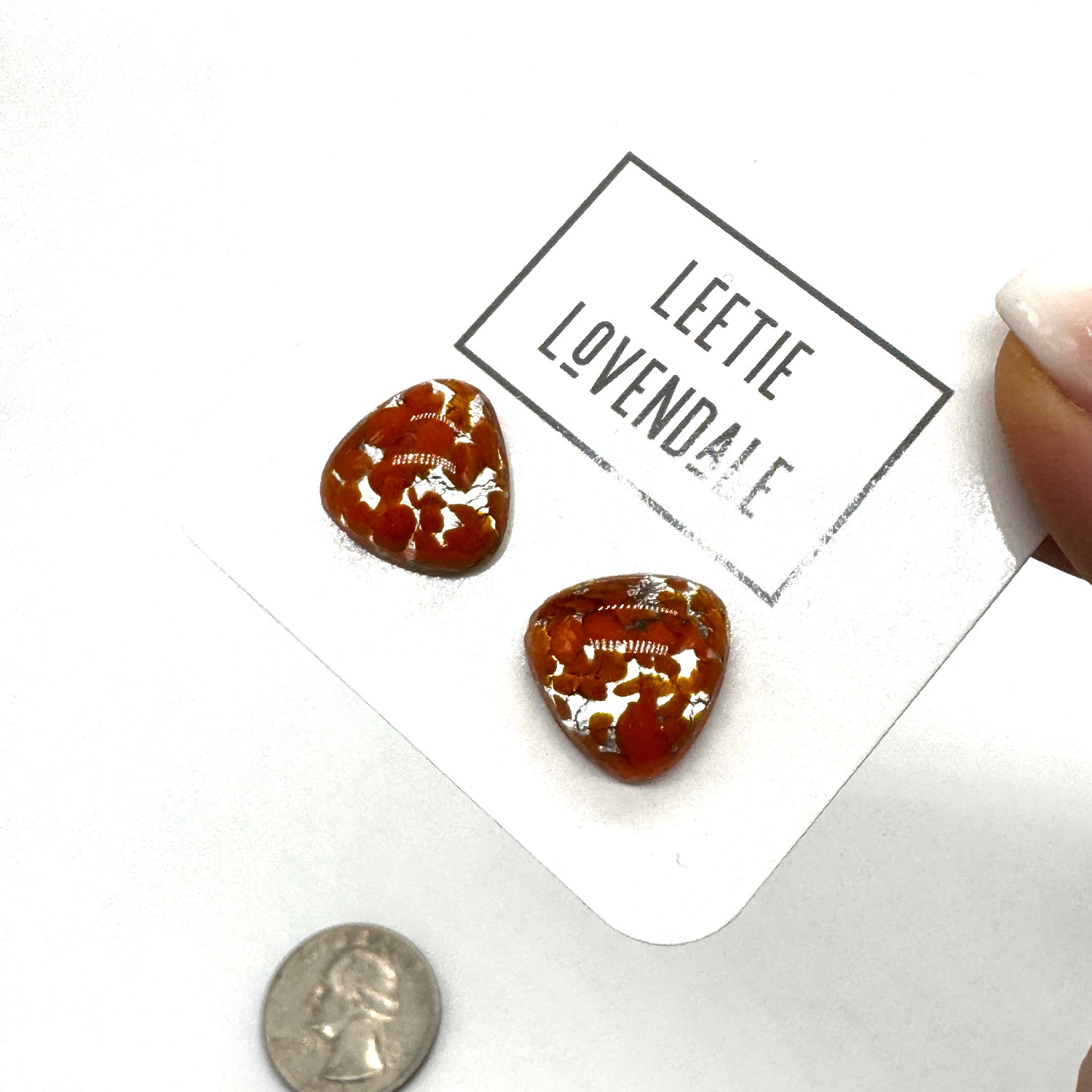 Two small triangular-shaped clear glass stud earrings with orange inclusions, displayed on a white card printed with the name &