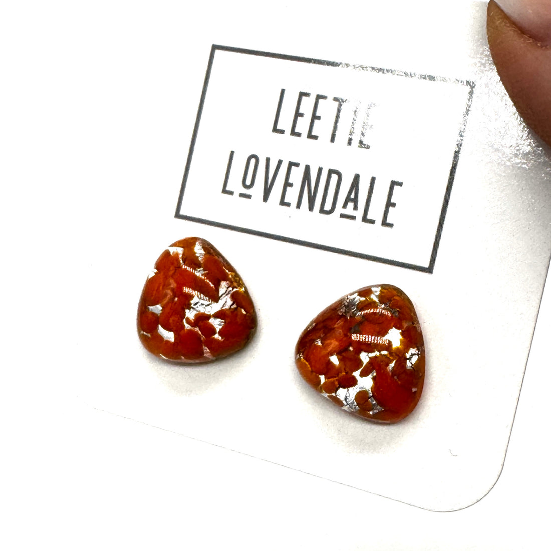 Two small triangular-shaped clear glass stud earrings with orange inclusions, displayed on a white card printed with the name &