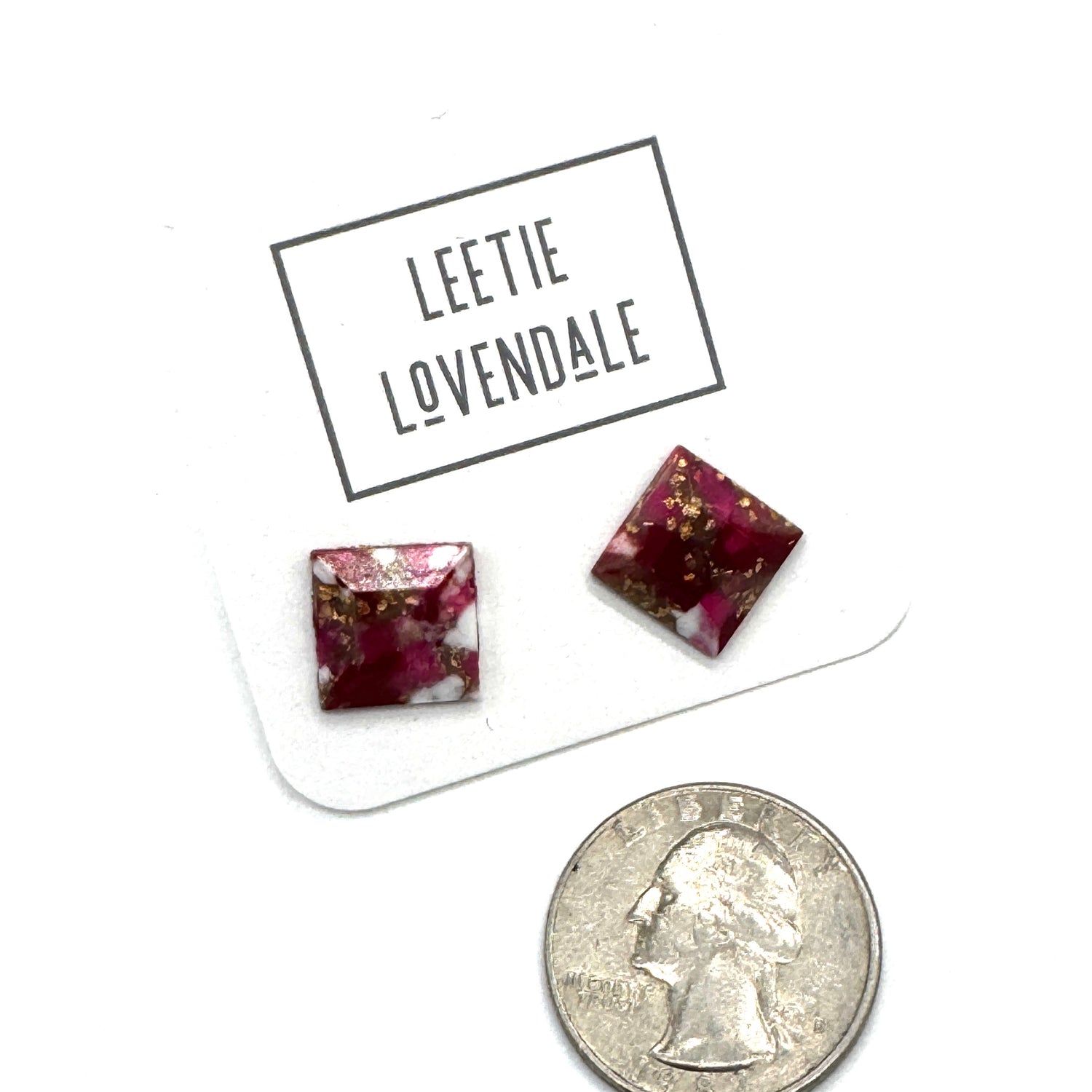 A pair of square glass stud earrings with a deep magenta-pink and white marbled pattern and gold flecks, displayed on a white card labeled &