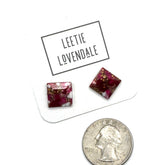 A pair of square glass stud earrings with a deep magenta-pink and white marbled pattern and gold flecks, displayed on a white card labeled &