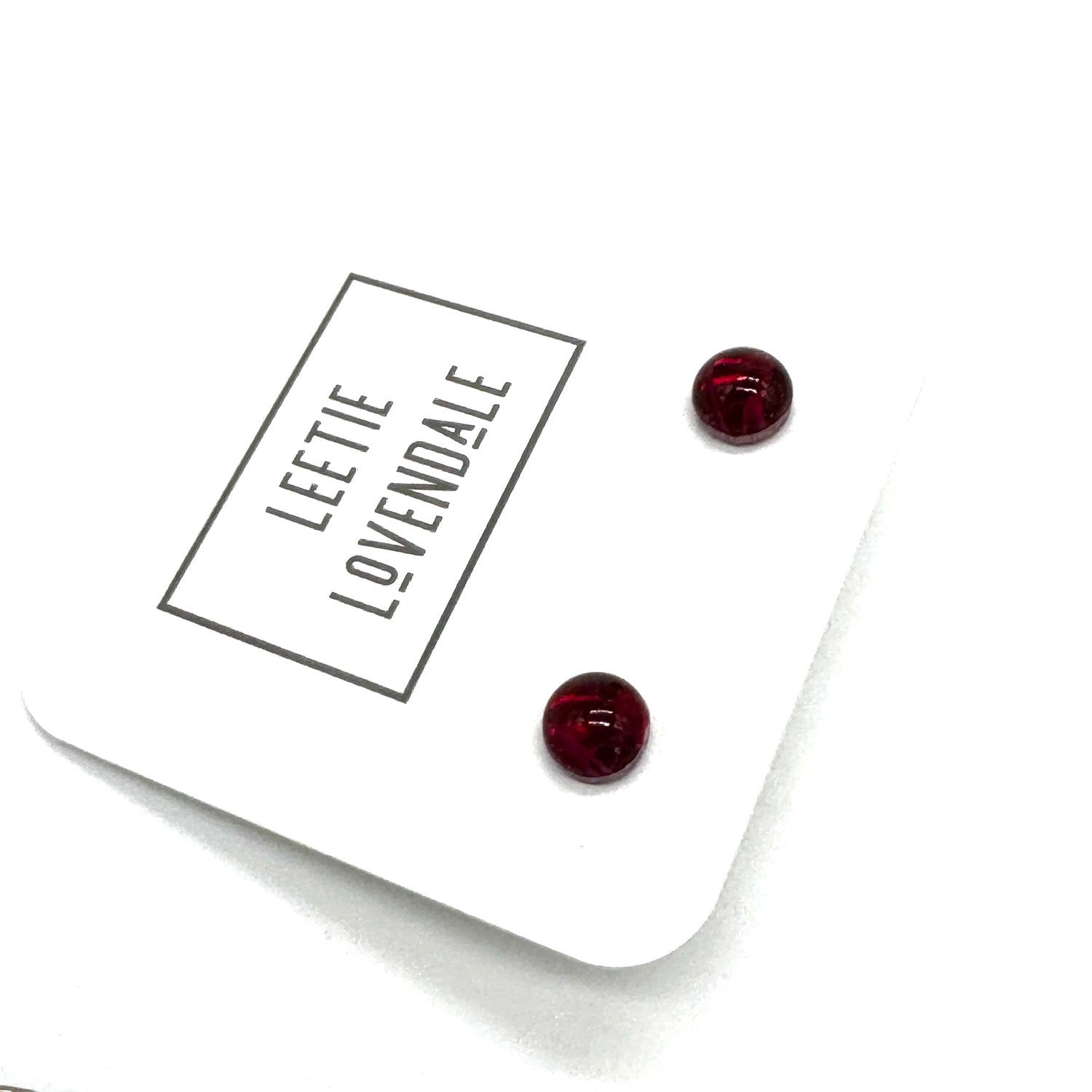 A pair of small round stud earrings in deep red resin, displayed on a white branded card that reads &