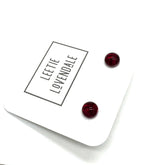 A pair of small round stud earrings in deep red resin, displayed on a white branded card that reads &