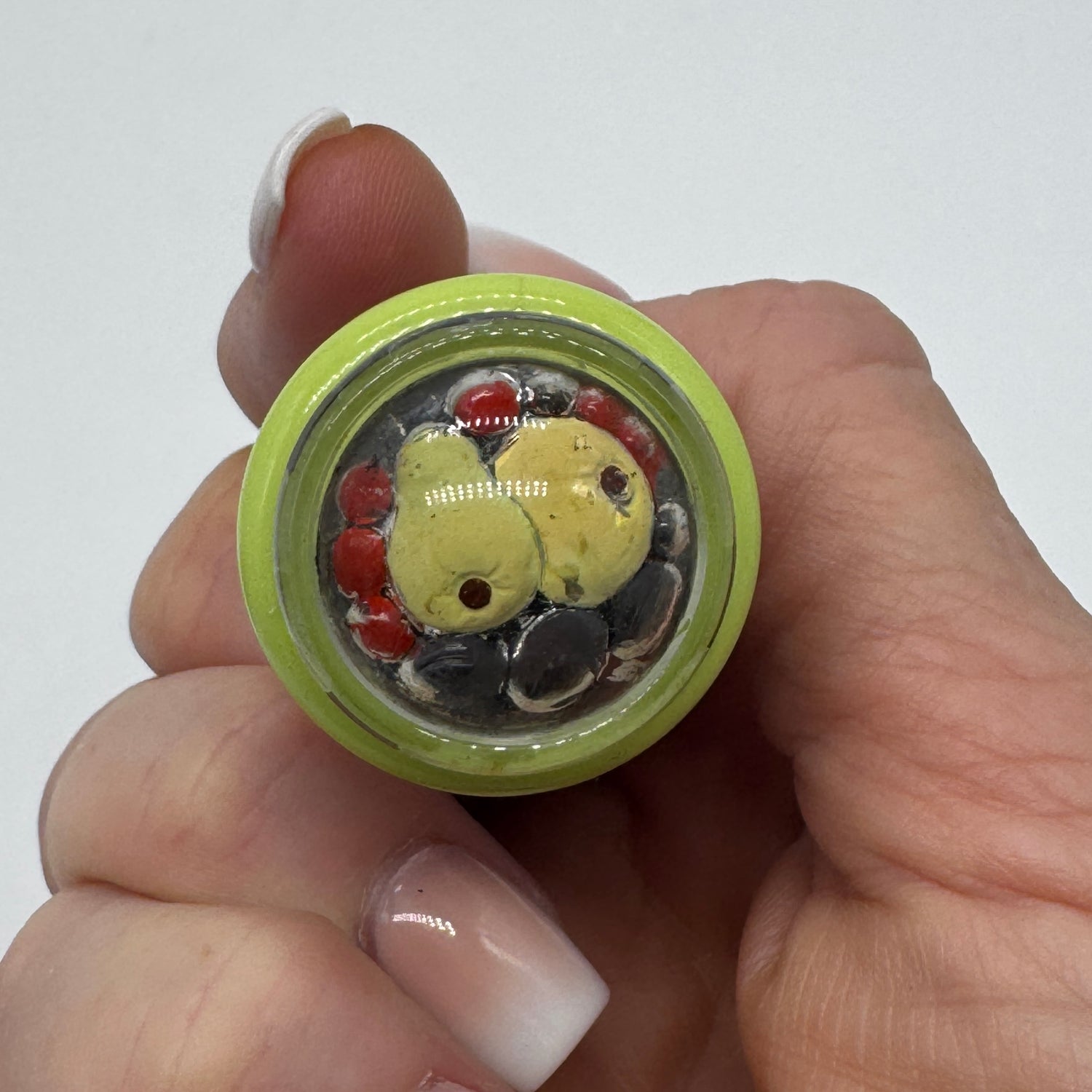 A hand holding a small round glass ring with a light green rim and a colorful interior featuring yellow, red, black, and white glass pieces under a clear top.