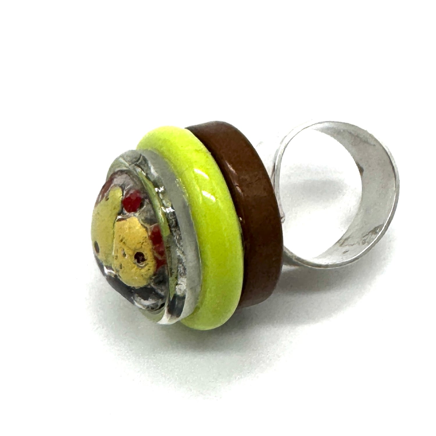 A handmade glass ring featuring a silver-toned band, a dark brown band, a bright lime green band, and a round clear glass cabochon containing a yellow bear face with red details.