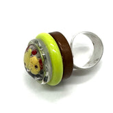 A handmade glass ring featuring a silver-toned band, a dark brown band, a bright lime green band, and a round clear glass cabochon containing a yellow bear face with red details.