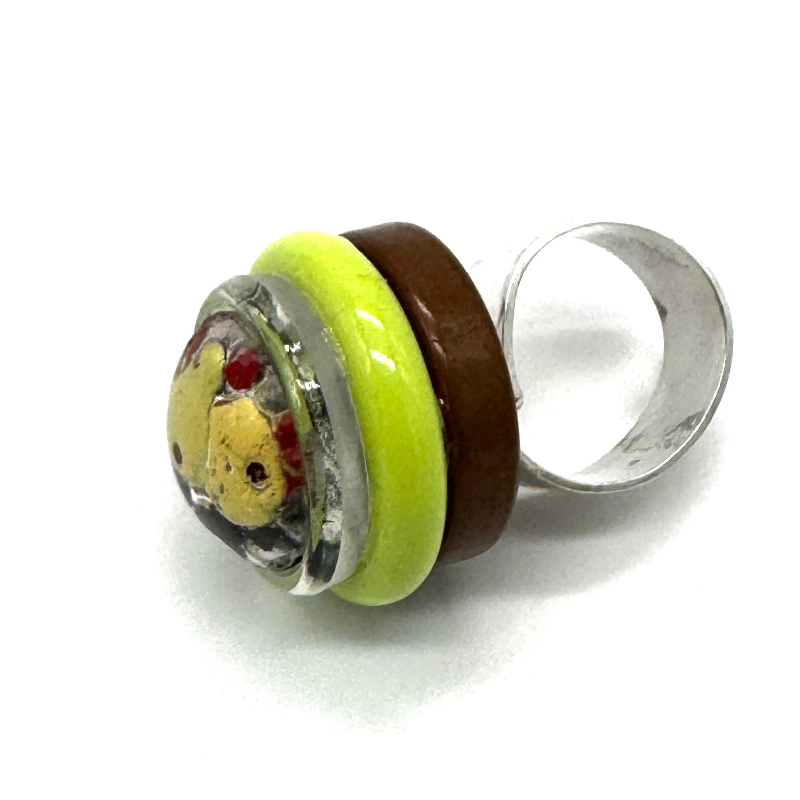 A handmade glass ring featuring a silver-toned band, a dark brown band, a bright lime green band, and a round clear glass cabochon containing a yellow bear face with red details.