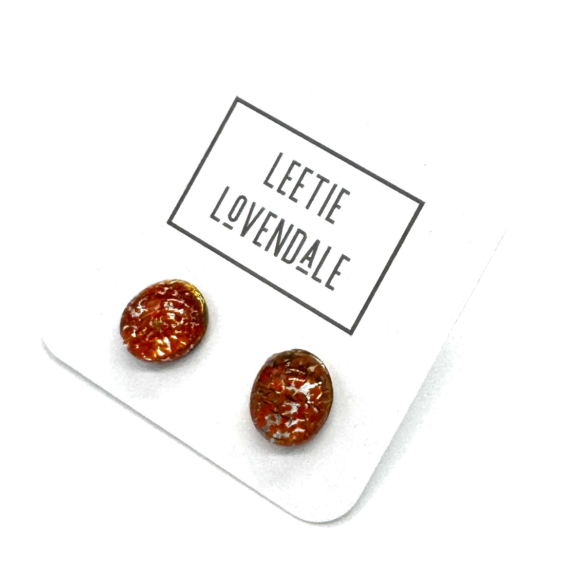 A pair of oval stud earrings with a marbled orange pattern, displayed on a white card printed with the brand name &