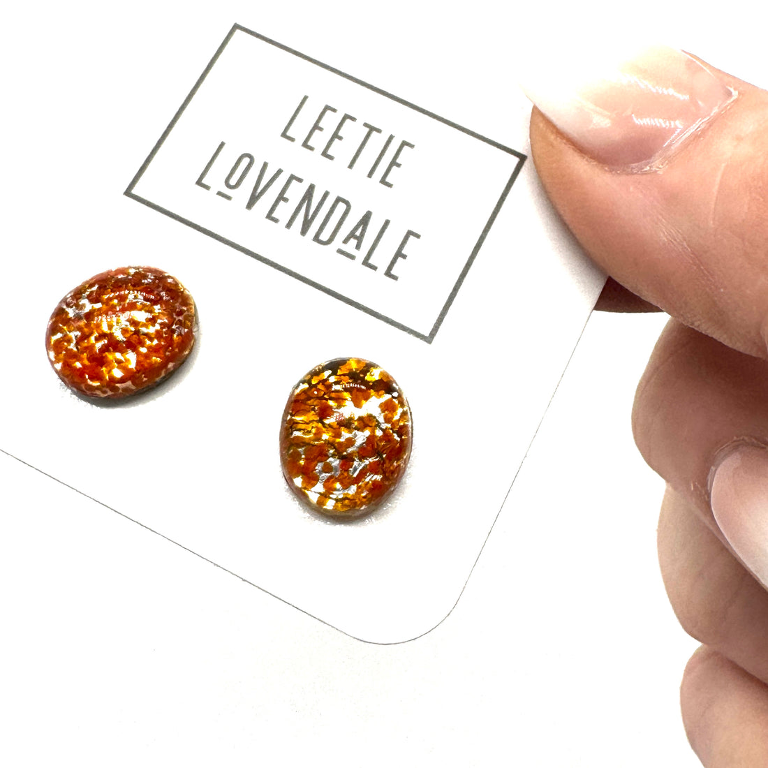 A close-up of a hand holding a white display card with two oval stud earrings. The card is branded &