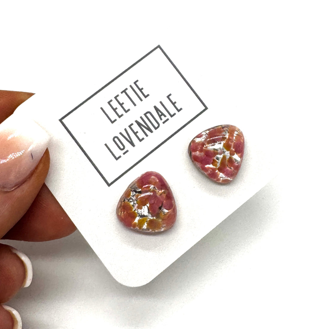 A close-up of a hand holding a white display card with two small triangular stud earrings. The card is branded &