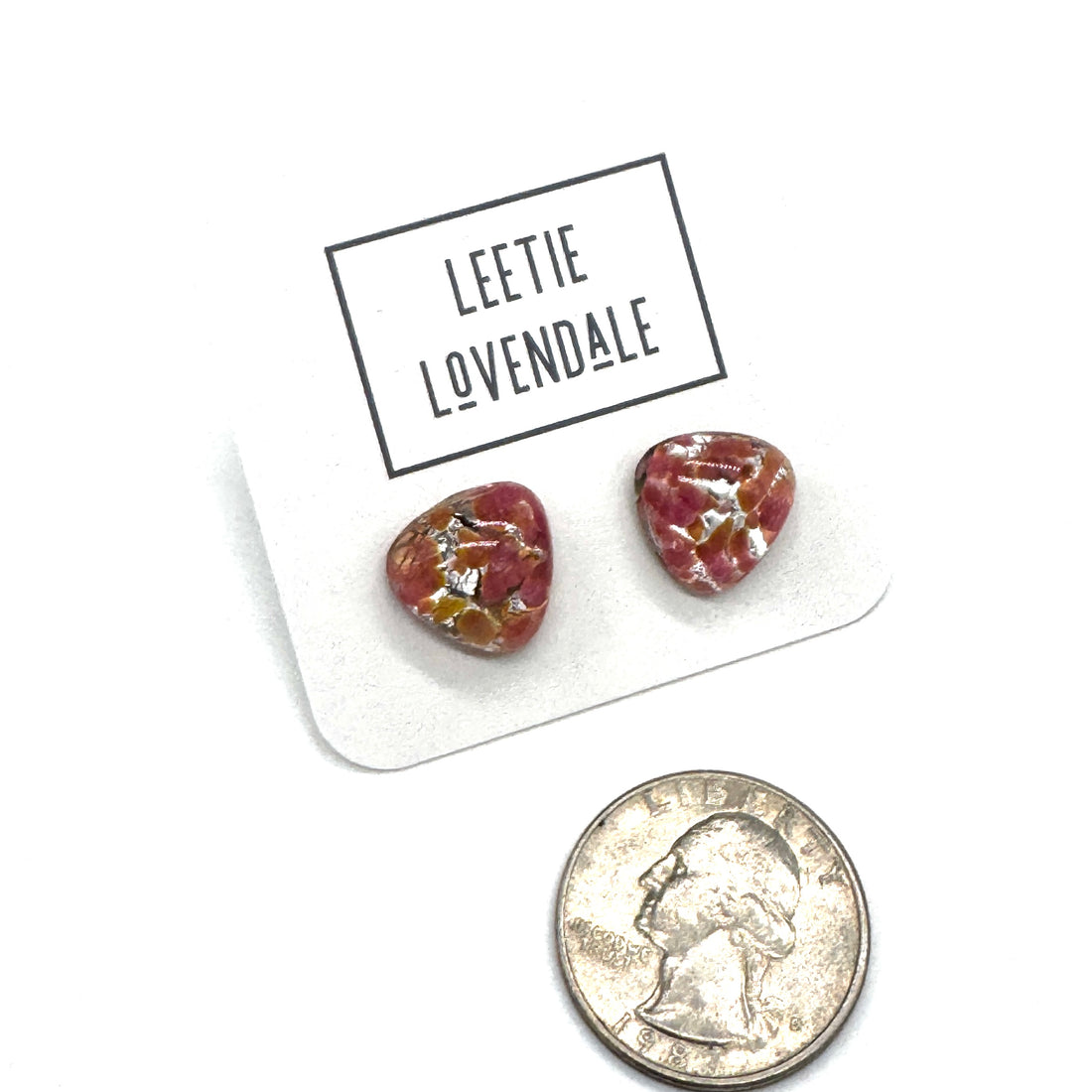 A pair of small irregular-shaped stud earrings with a marbled pink and gold pattern, displayed on a white card labeled &