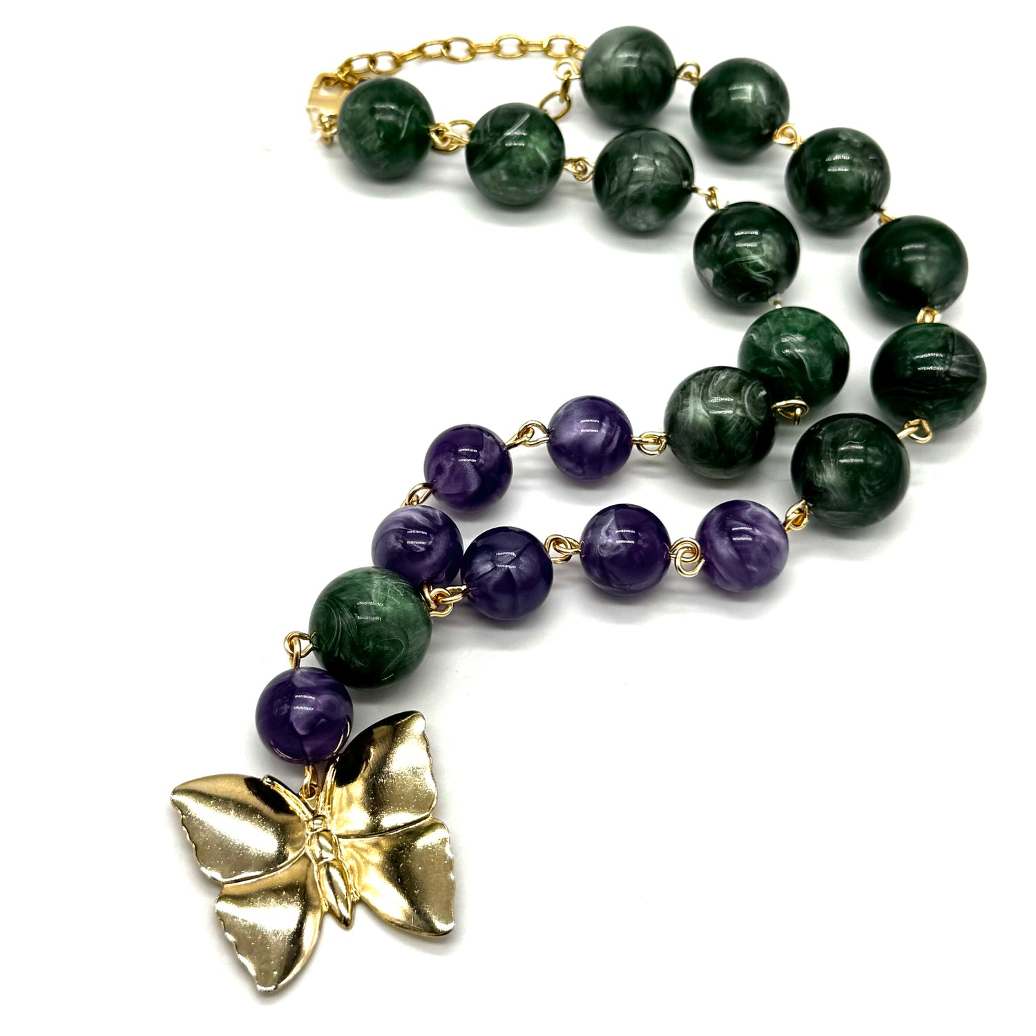 A beaded necklace featuring dark green lucite beads, purple amethyst beads, and a textured gold butterfly pendant, displayed on a clean white background.