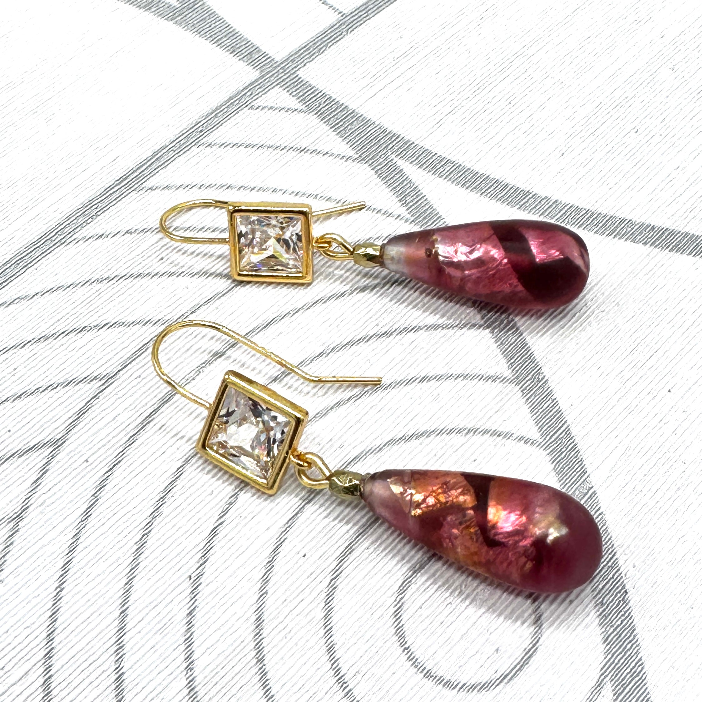A pair of gold-tone dangle earrings with square clear crystal settings and teardrop-shaped burgundy glass drops, displayed on a white textured background with gray circular line patterns.
