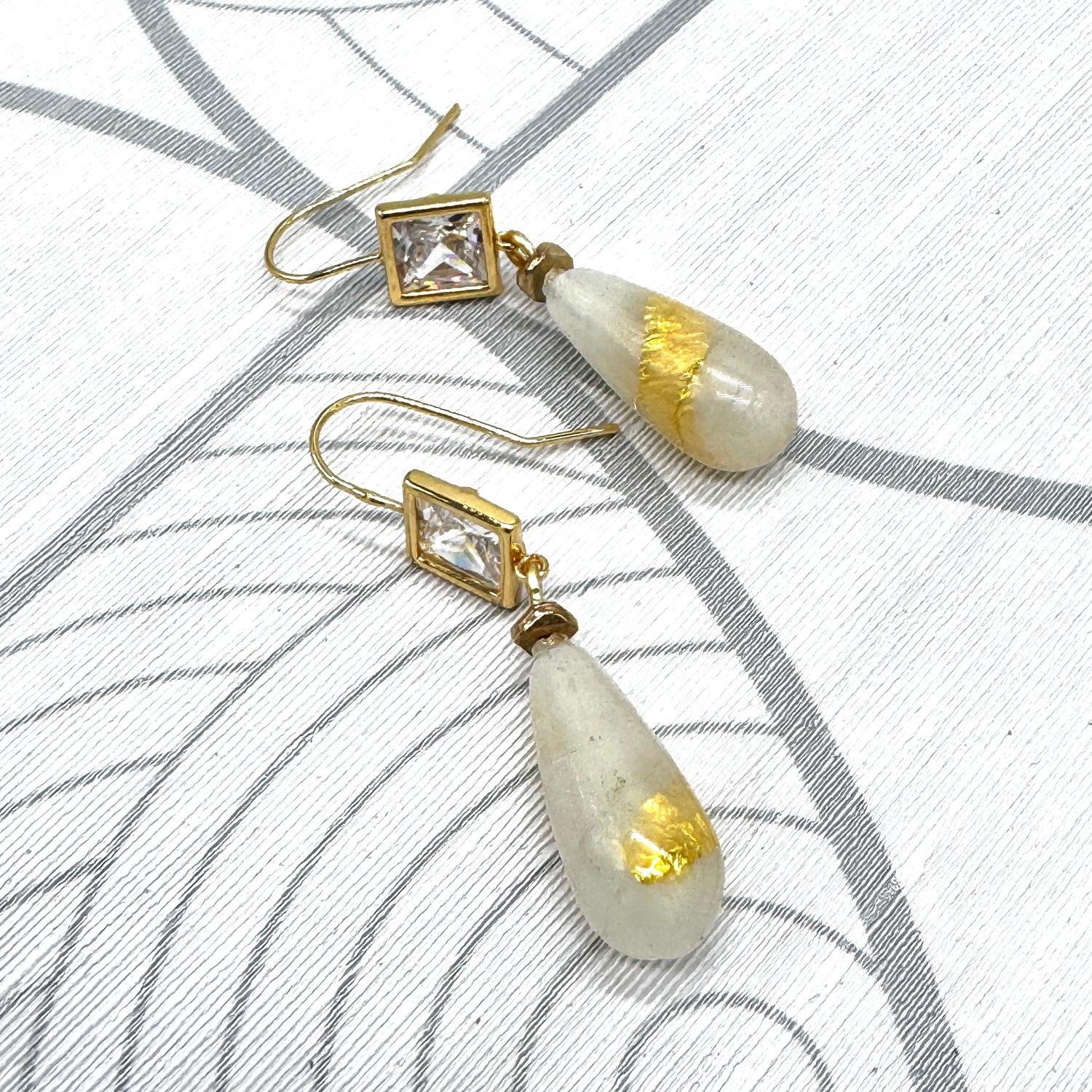 A pair of gold-tone dangle earrings with clear square-cut stones and teardrop-shaped milky white beads containing gold leaf inclusions, displayed on a white textured surface with gray curved line patterns.