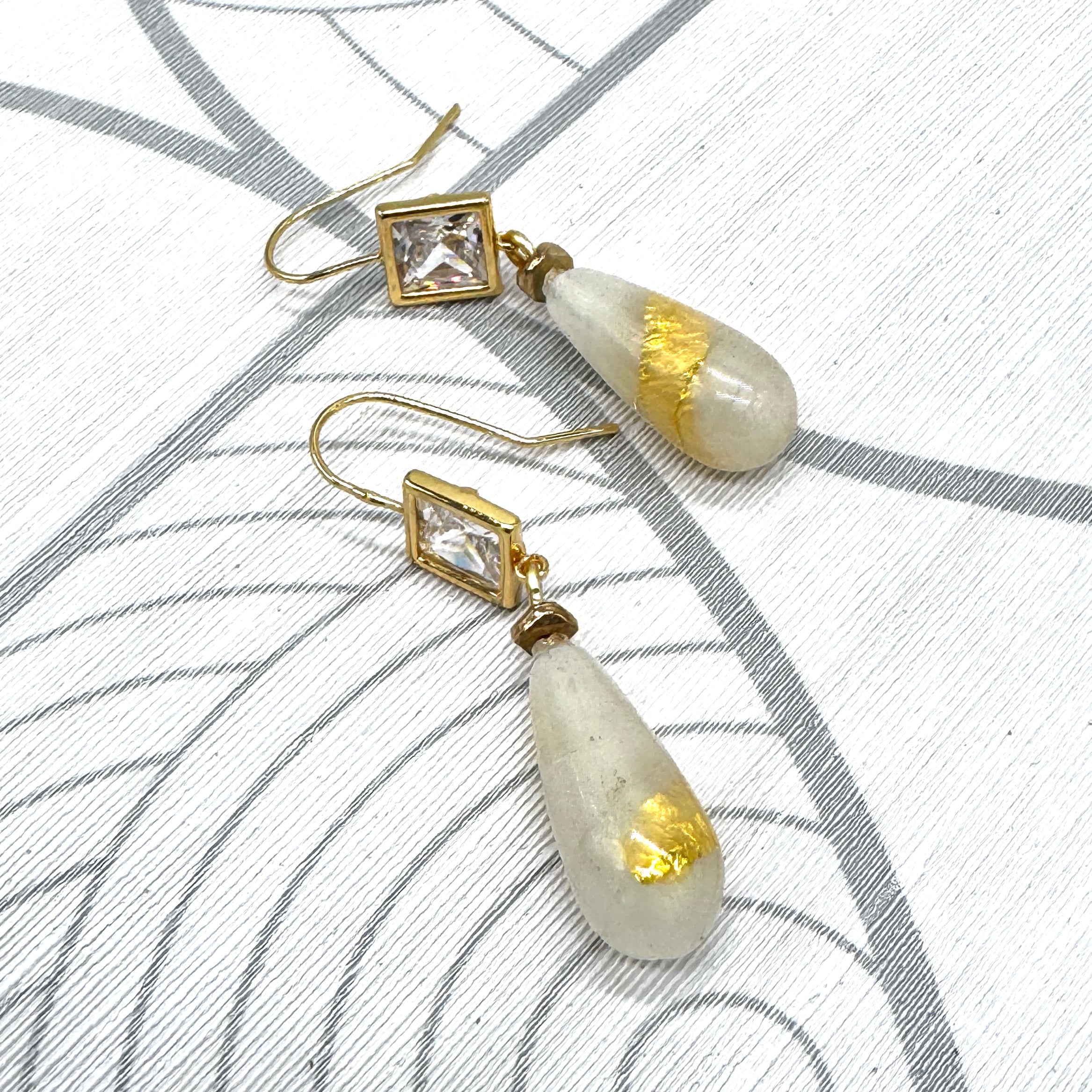 A pair of gold-tone dangle earrings with clear square-cut stones and teardrop-shaped milky white beads containing gold leaf inclusions, displayed on a white textured surface with gray curved line patterns.