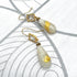 A pair of gold-tone dangle earrings with clear square-cut stones and teardrop-shaped milky white beads containing gold leaf inclusions, displayed on a white textured surface with gray curved line patterns.