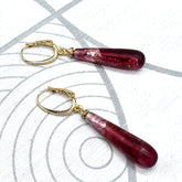 A pair of dangling earrings with teardrop-shaped deep burgundy glass beads and gold leverback clasps, displayed on a white textured surface with gray concentric circle patterns.