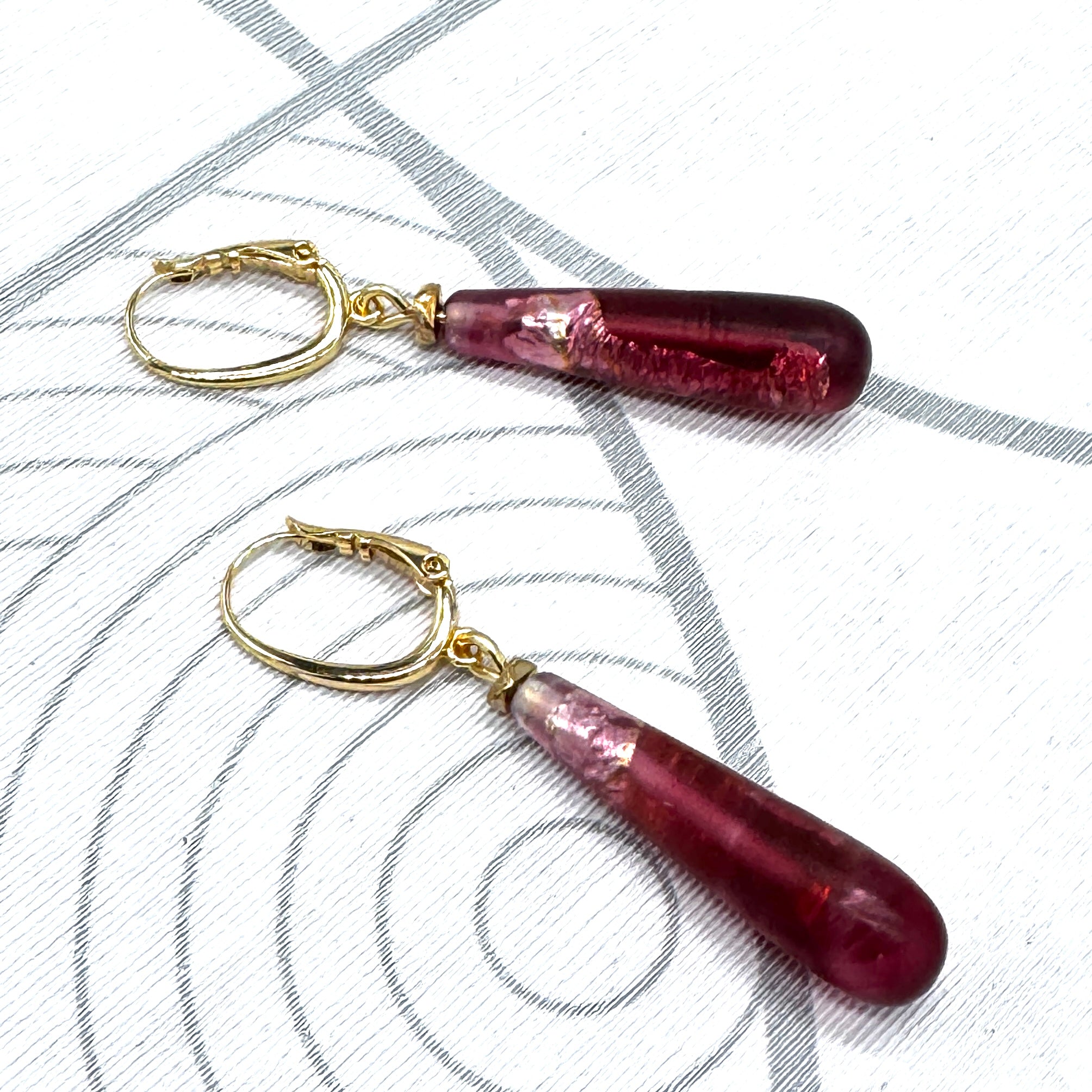 A pair of dangling earrings with teardrop-shaped deep burgundy glass beads and gold leverback clasps, displayed on a white textured surface with gray concentric circle patterns.