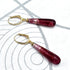 A pair of dangling earrings with teardrop-shaped deep burgundy glass beads and gold leverback clasps, displayed on a white textured surface with gray concentric circle patterns.