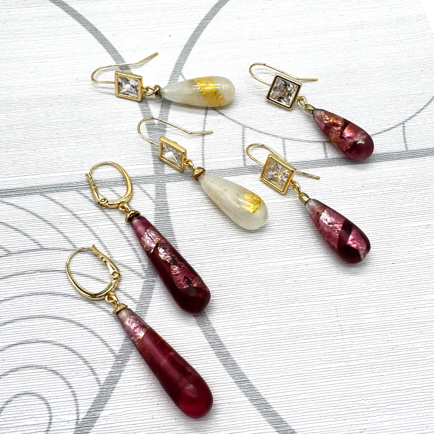 Three pairs of handmade drop earrings arranged on a light gray patterned background. The earrings feature gold-tone metal settings with clear crystals and teardrop-shaped glass drops in deep burgundy and milky white.