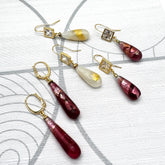 Three pairs of handmade drop earrings arranged on a light gray patterned background. The earrings feature gold-tone metal settings with clear crystals and teardrop-shaped glass drops in deep burgundy and milky white.